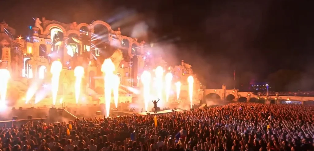 Illustration de Komby Dot IO face à une foule dense, avec une scène de festival et des colonnes de feu.